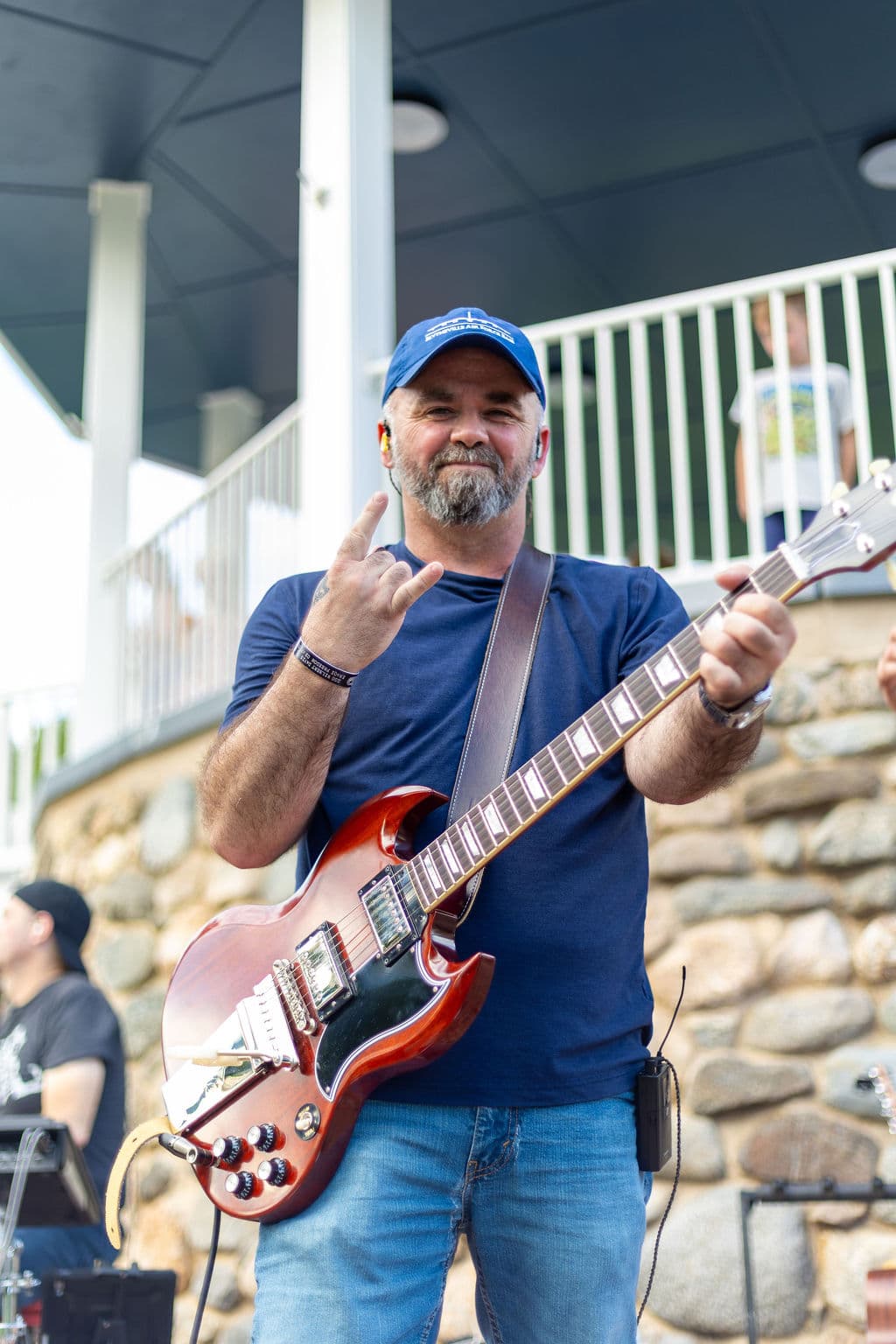 Guitarist giving a rock-on hand sign on an outdoor bandstand with onlookers, including a kid, on the porch behind him
